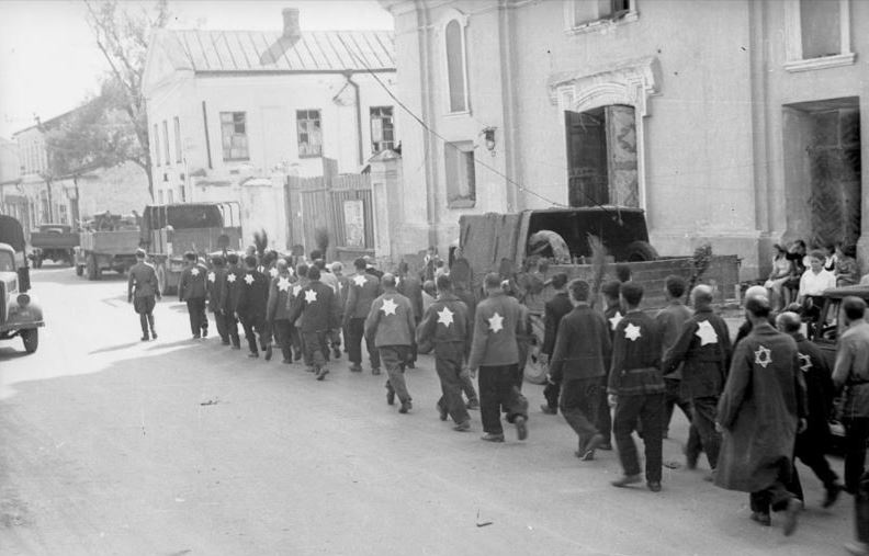 Eine Gruppe von Männern geht am Straßenrand entlang. Auf der Kleidung tragen die Männer helle Sterne. Vorne geht ein Soldat in Uniform. Rechts im Bild stehen einige Passant:innen. Die Straße ist von Häusern gesäumt.  © Wikimedia, Bundesarchiv, Bild 101I-138-1083-31, Rudolf Kessler, Kolonne von Juden mit aufgenähten Judenstern und Spaten unter Bewachung von Wehrmachtssoldaten beim Marsch durch die Stadt, CC-BY-SA 3.0. 