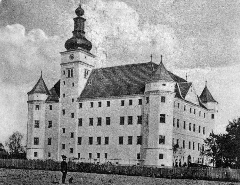 Die Postkarte in schwarz-weiß zeigt ein Schloss mit hellen Außenmauern und Reihen kleiner dunkler Fenster. An den Ecken befinden sich kleine Türmchen. Auf einer Seite hat das Schloss einen großen Turm mit Uhr. Vor dem Schloss steht ein Mann mit einem Hund auf einem Feld. © Wikimedia, Schloss Hartheim, 1924