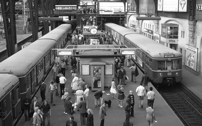 Auf dem Bahnsteig tummeln sich Menschen, rechts und links stehen auf den Gleisen Züge. In der Mitte des Bahnsteigs sind kleine Warteräume, Anzeigetafeln und eine Uhr. © Wikimedia