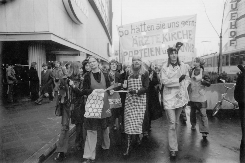 Ein Demonstrationszug, an dessen Spitze vier Frauen marschieren, eine davon mit Besen in der Hand, zwei mit Topfdeckel und Kochlöffel. Dahinter ein Banner mit der Aufschrift „So hätten sie uns gern: Ärzte, Kirche, Parteien und Männer“. Links sind Passanten vor einem Gebäude zu sehen.  © Wikimedia, Inge Werth, Frauenprotest gegen Paragraph 218, Frankfurt 1974 (CC BY-SA 4.0). 