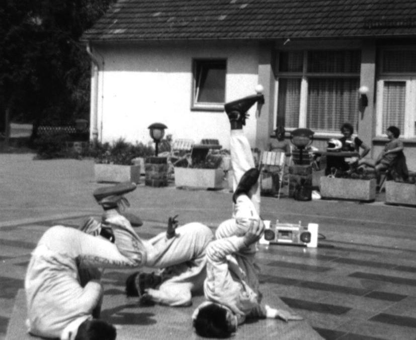 Drei Jugendliche liegen auf einer Matte und machen Breakdance. Die Beine haben sie in der Luft. Sie tragen weiße Sportanzüge. Im Hintergrund steht ein Haus mit Blumenkästen davor. Ein Kassettenrekorder steht am Boden.  © Privatsammlung Heiko Hahnewald.