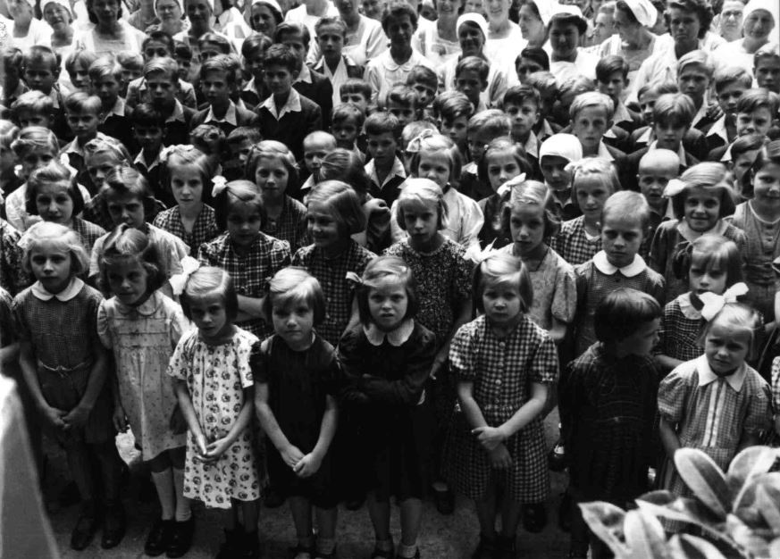 Zahlreiche Kinder stehen in einer Gruppe beieinander und schauen zur fotografierenden Person hoch. Vorne stehen in mehreren Reihen Mädchen in Kleidern, dahinter Buben. In den hintersten Reihen stehen Erwachsene in weißen Kitteln und mit weißen Hauben.  © ÖNB Bildarchiv, Kinder am Steinhof nach ihrer Befreiung 1945. 