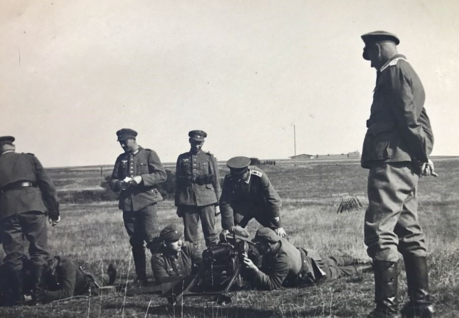 Mehrere Soldaten stehen auf einem Feld. Vier liegen am Boden und hantieren mit Kriegsgerät. Die Männer tragen Uniformen und Kappen der Wehrmacht.  © Wikimedia, Paul Calwer, Ausbildung von Wehrmachtssoldaten bei Regensburg (CC-BY-SA-4.0). 