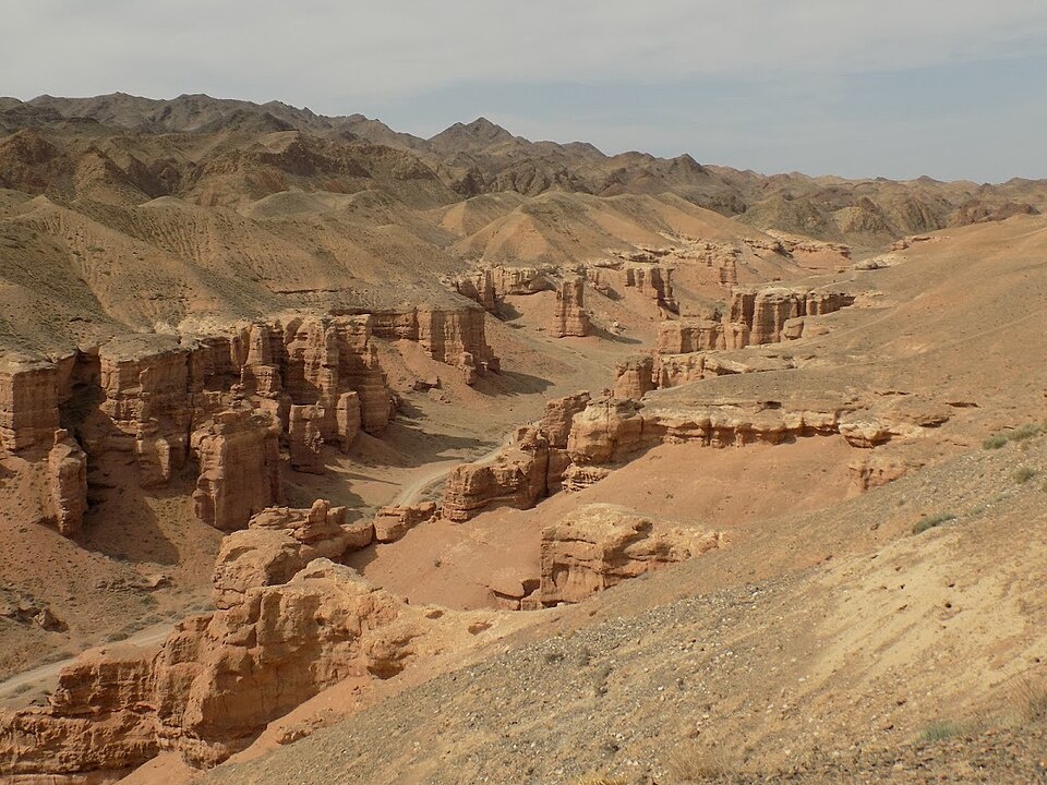 Das Foto zeigt einen Canyon mit Bergen im Hintergrund vor einem graublauen Himmel. Die Erde und das Gestein des Canyons sind orange.  © Wikimedia, Anna Dobrynina, Canyon in Kazakhstan, 2017 (CC-BY-3.0).  